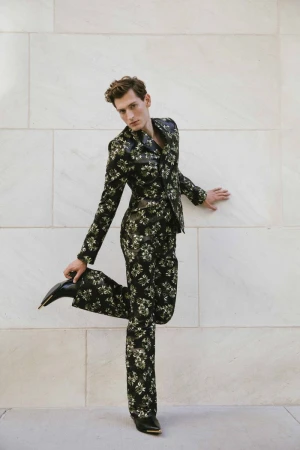 Male fashion model strikes a pose in a floral suit in front of a neutral wall, showcasing fashion style and visual appeal.