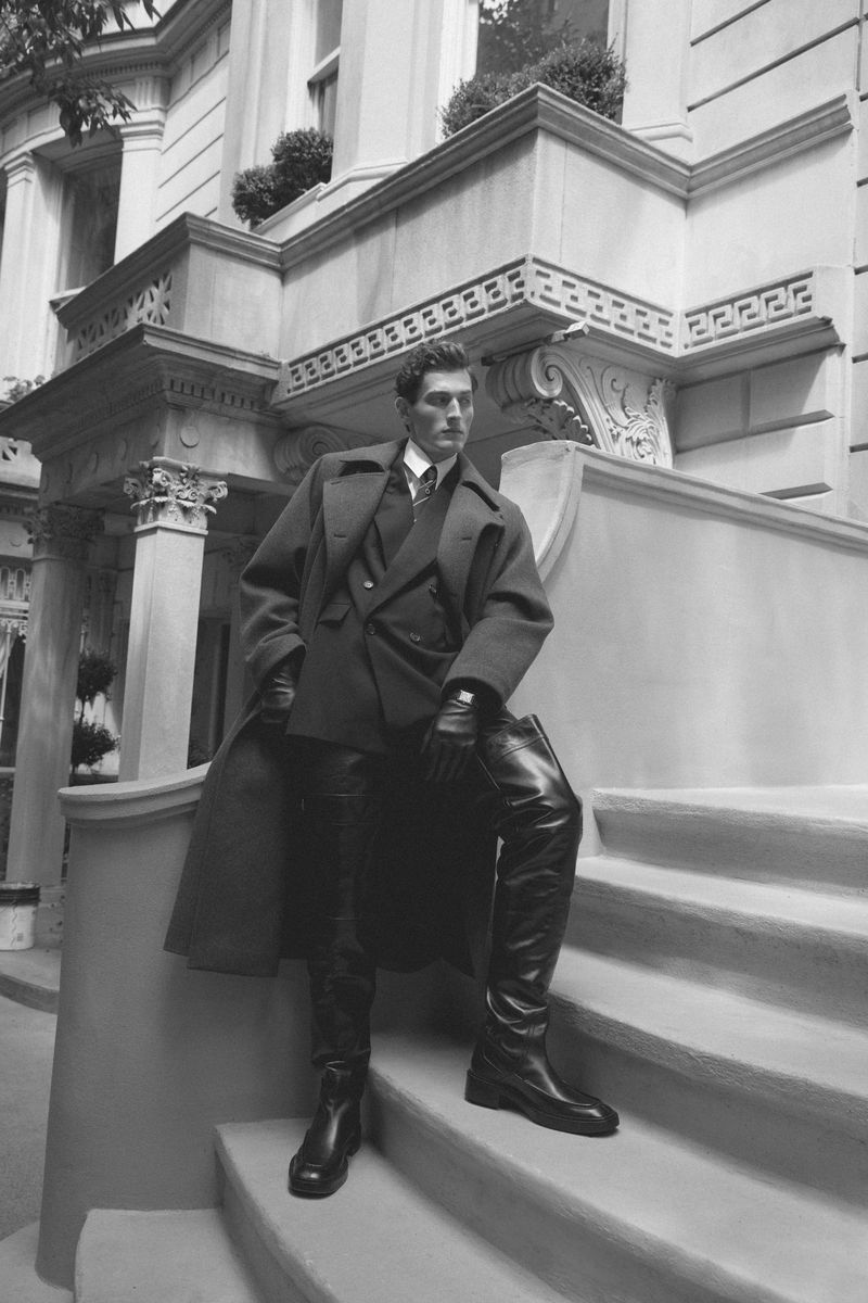 Man in overcoat and boots poses on steps of urban brownstone building in a black and white fashion photograph.