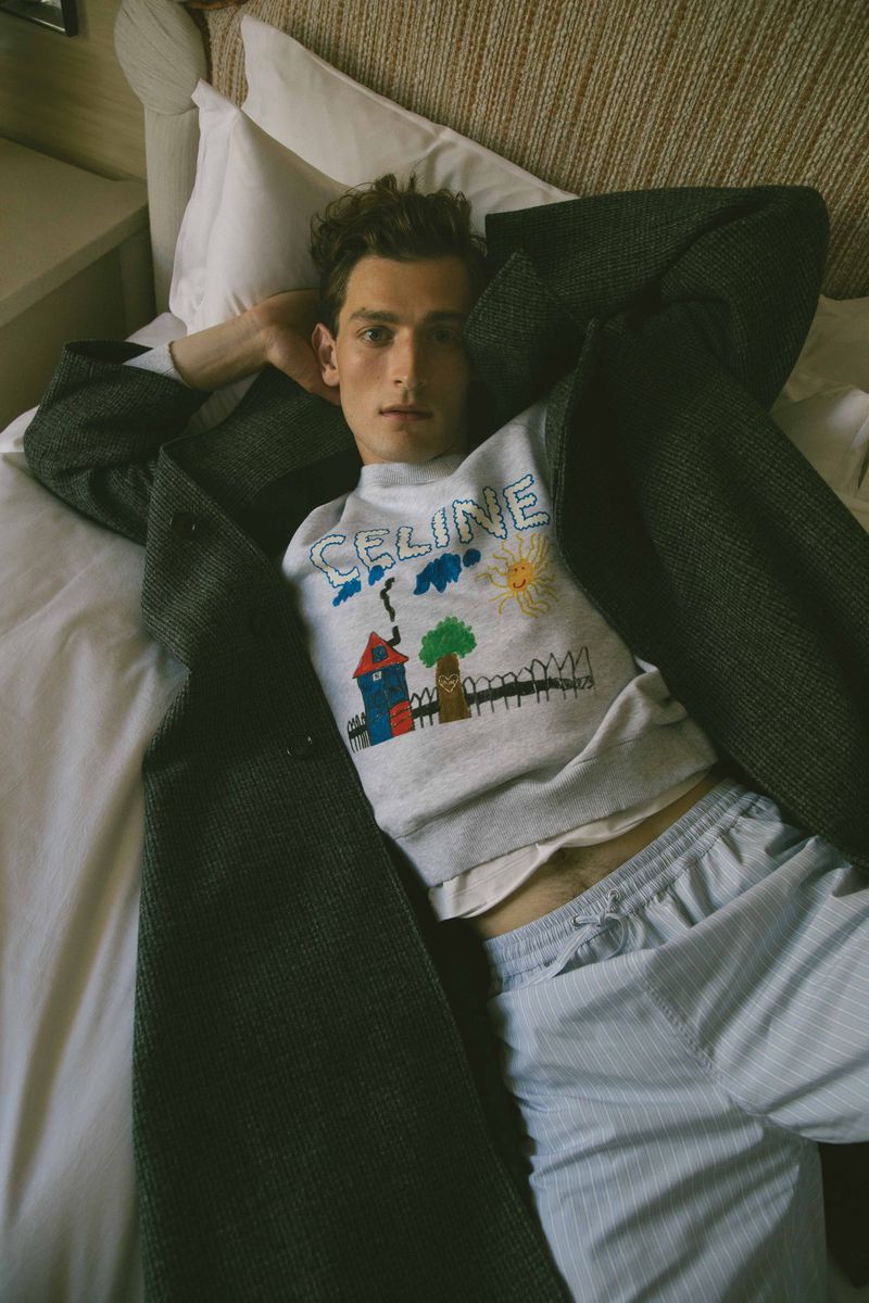 A fair-skinned young man lies on a bed with a grey coat on top of a white sweatshirt featuring a colorful drawing and the Celine logo.
