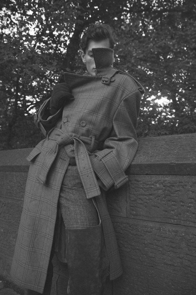 Monochrome portrait of a stylish man in a trench coat, set against a blurred tree background, exuding a vintage fashion editorial aesthetic.