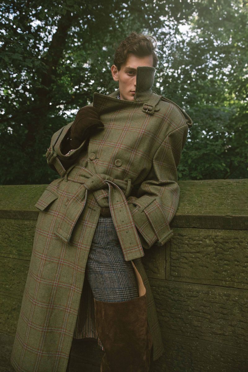 Fashionable adult male in green plaid trench coat poses against a wall in an outdoor setting with green foliage in the background.
