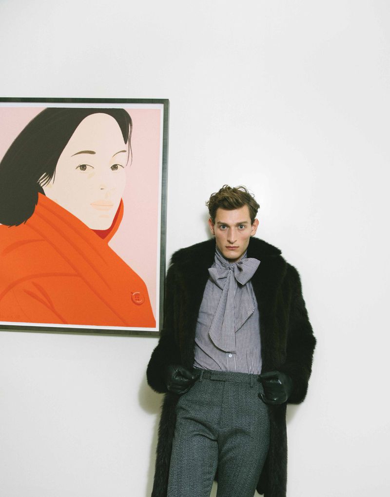 A young adult male with dark hair in a fur coat and gray pants, posing indoors next to a framed portrait with an off-white background.