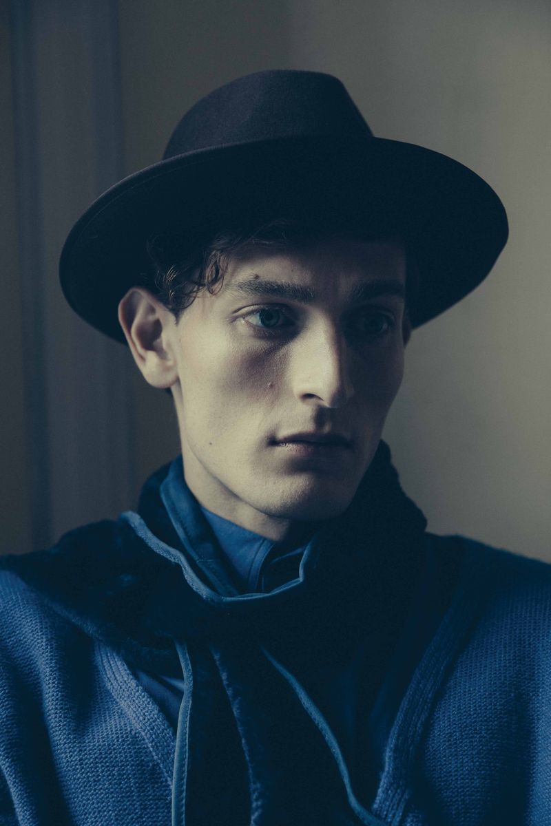 Headshot of a young adult male wearing a fedora and blue shirt and sweater against a neutral indoor backdrop in soft lighting.
