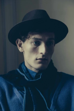 Headshot of a young adult male wearing a fedora and blue shirt and sweater against a neutral indoor backdrop in soft lighting.