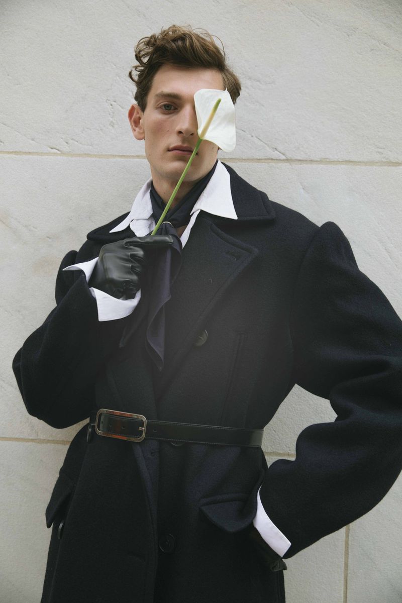 Adult male model in a black overcoat poses with a white flower against a neutral background, wearing gloves and a button-down shirt.