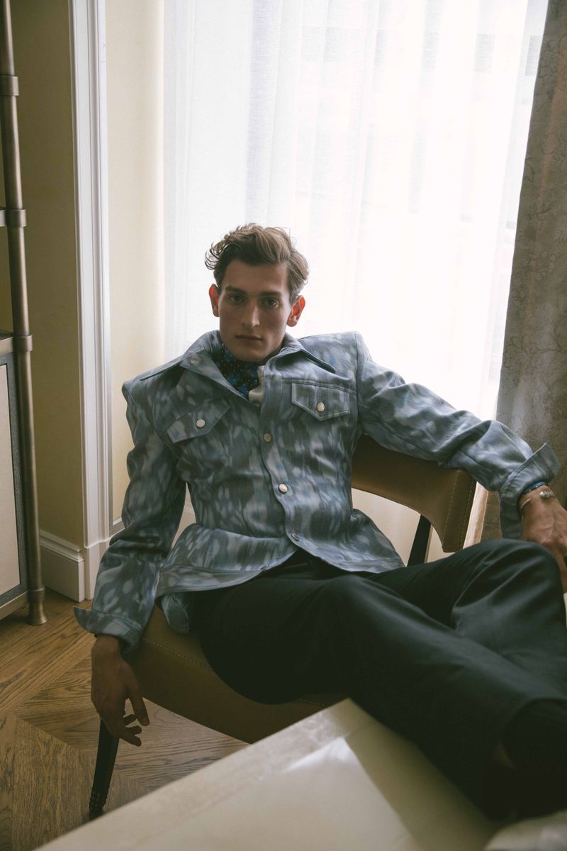 Adult male in a patterned jacket poses while seated in a chair in a room with hardwood floors and natural light from a window.
