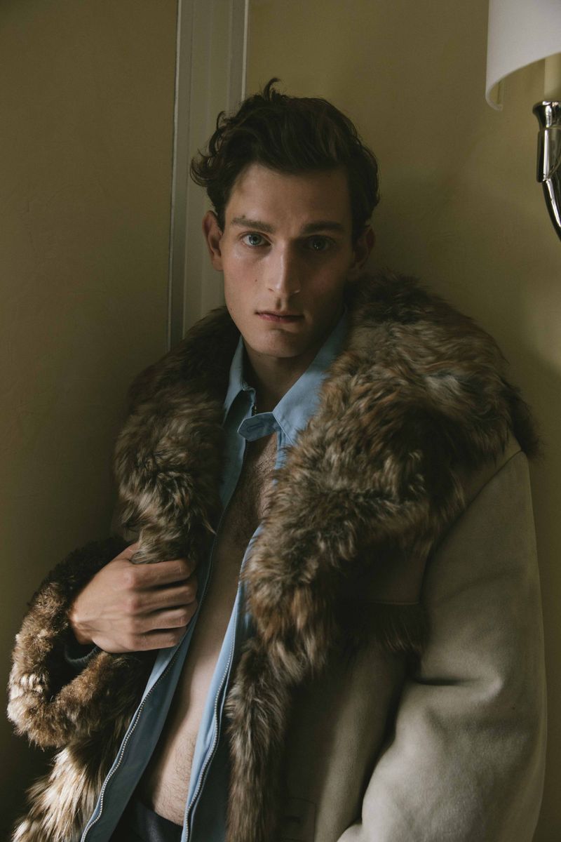 Portrait of a young adult male with dark curly hair, wearing a light blue shirt and a fur-lined coat, posing indoors against a neutral wall.