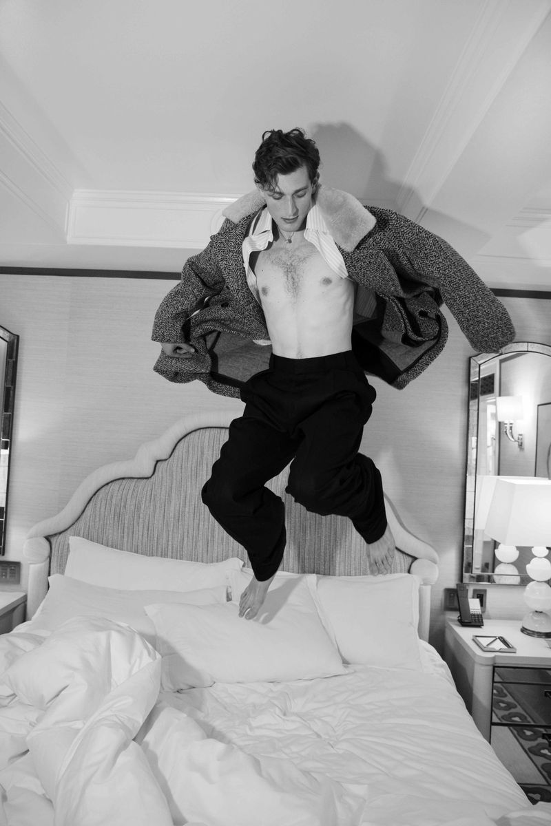 Black and white portrait of a happy adult male jumping barefoot on a bed in a bedroom. He is wearing a coat and dark pants.