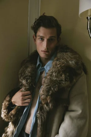 Portrait of a man wearing a light blue unbuttoned shirt and a heavy fur-trimmed coat leaning against a warm interior wall.