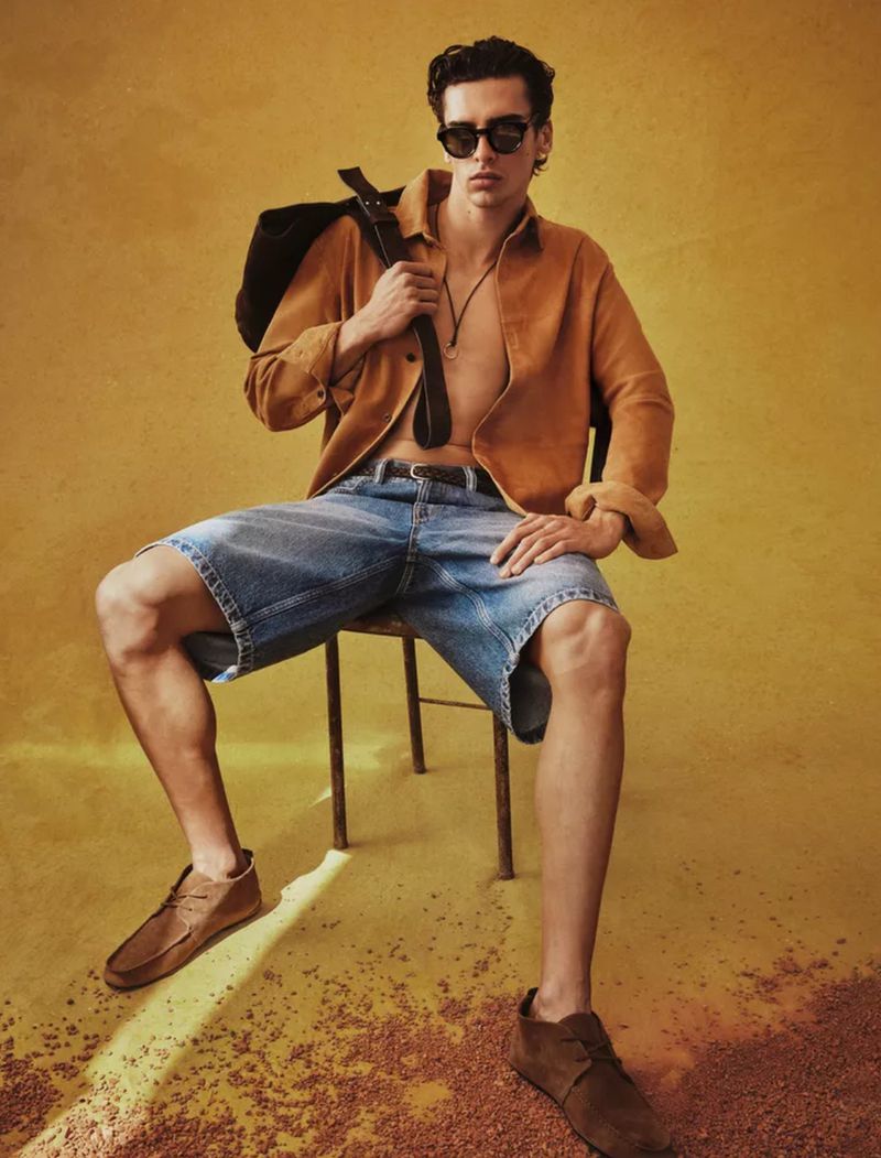 A stylish adult male sits on a metal chair wearing blue jean shorts, a tan suede shirt, sunglasses, and brown suede shoes in a studio.
