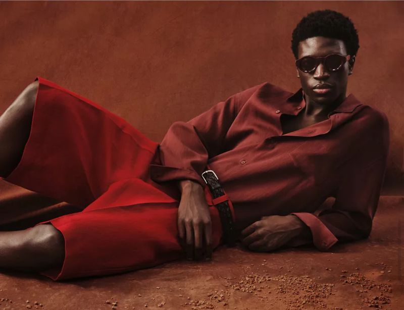 Model wearing dark sunglasses, a red shirt and shorts while reclining against a warm reddish-brown textured backdrop.