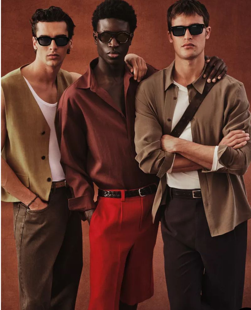 Three diverse young adult men pose in fashionable casual outfits and sunglasses against a solid warm-toned backdrop for a fashion shoot.