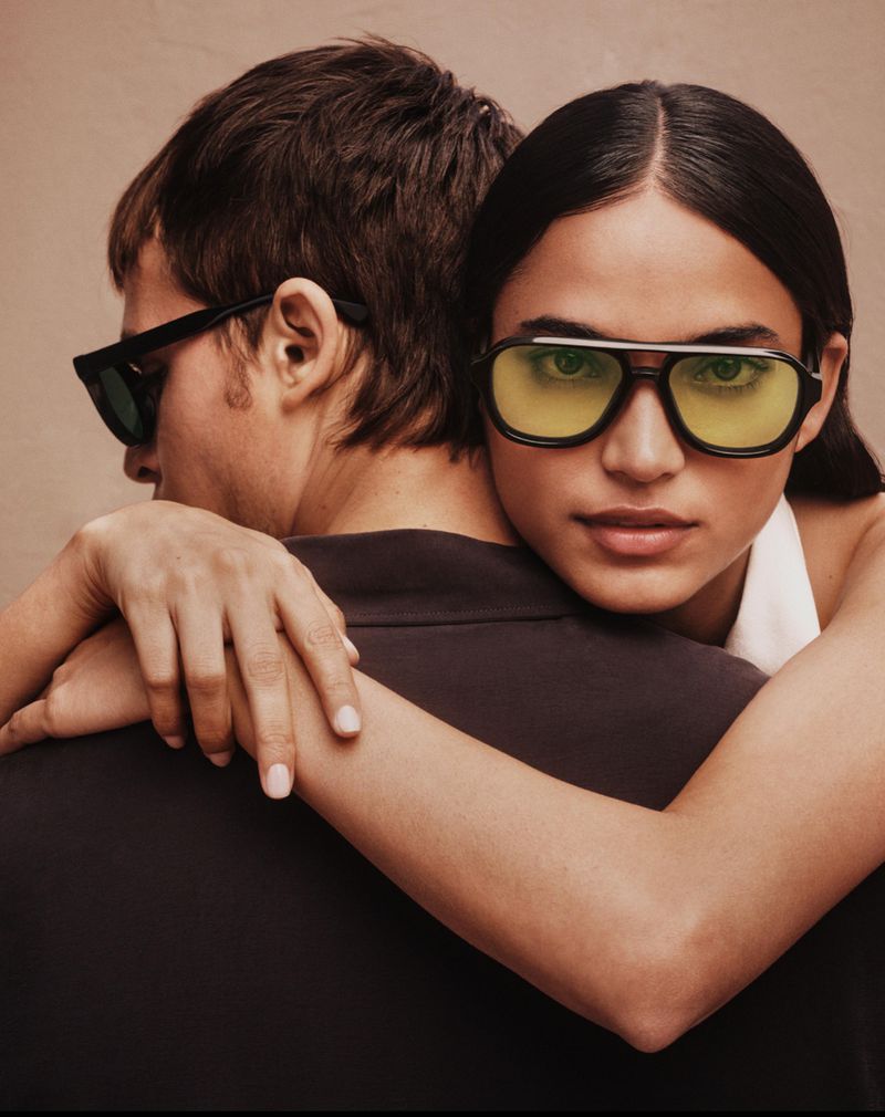 Fashion shot of a man and woman wearing sunglasses, the woman is embracing the man from behind with her arms around his shoulders.