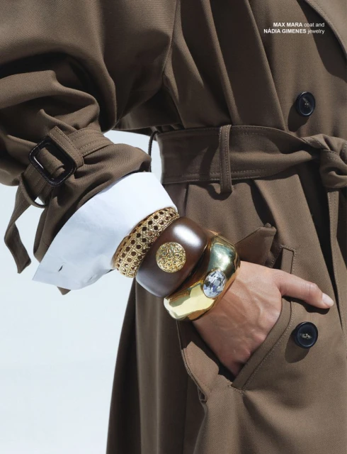 Fashion editorial shot showcases a hand adorned with stacked gold and wooden bracelets, against a neutral coat sleeve and white shirt cuff.