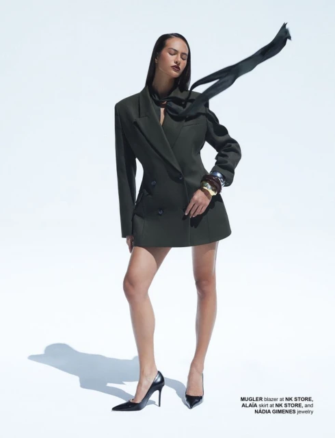 A fashion shot of an adult woman in a dark green blazer and skirt, accessorized with black heels and bold jewelry on a light studio backdrop.