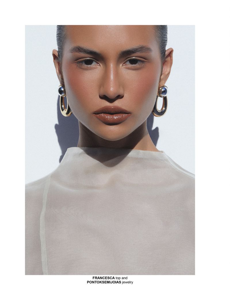 Close-up studio shot of a tan-skinned woman wearing statement gold earrings and a beige top against a white background with subtle shadows.