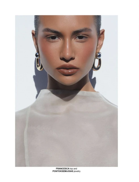 Close-up studio shot of a tan-skinned woman wearing statement gold earrings and a beige top against a white background with subtle shadows.