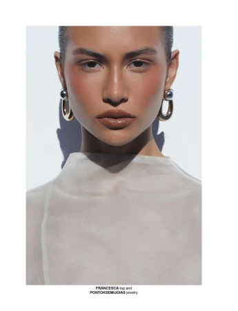 Close-up studio shot of a tan-skinned woman wearing statement gold earrings and a beige top against a white background with subtle shadows.