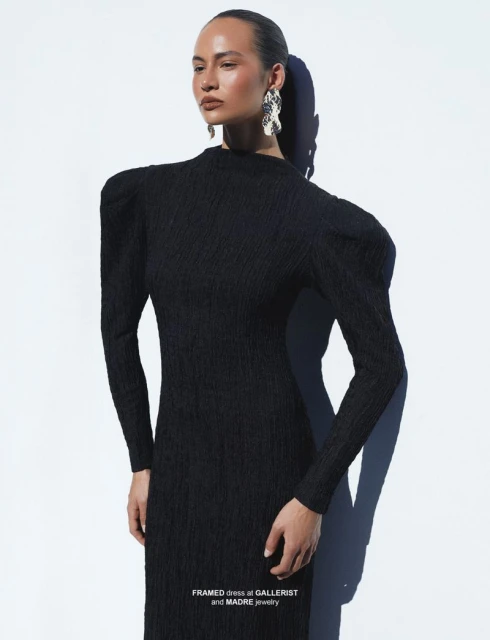 Elegant adult female wearing a textured black dress and ornate earrings poses against a bright white backdrop in a fashion editorial.