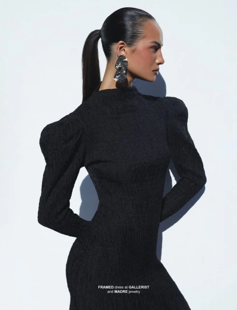 Woman poses in a textured black dress with statement earrings against a plain white background in a fashion photography style image.