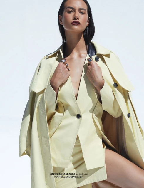 Fashionable woman in stylish yellow trench coat with plunging neckline, posing in front of a bright white backdrop, looking confident.