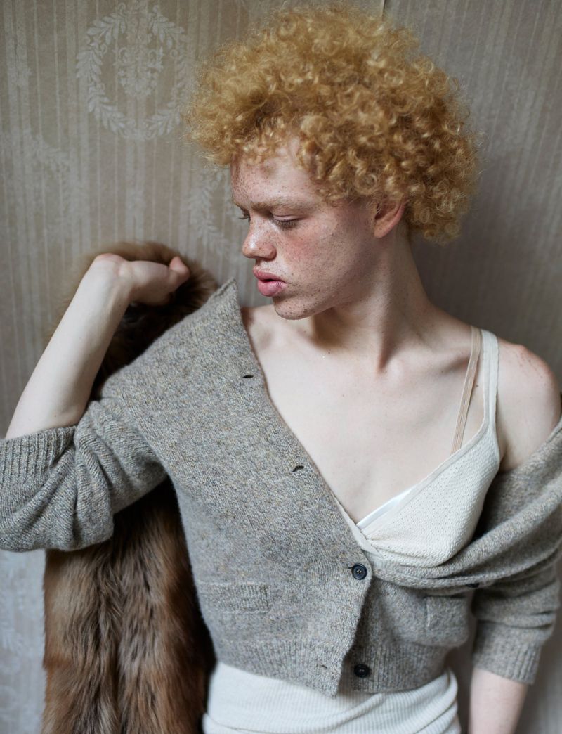 Portrait of a blonde adult with freckles wearing a cardigan and tank top in front of patterned wallpaper and a fur stole.