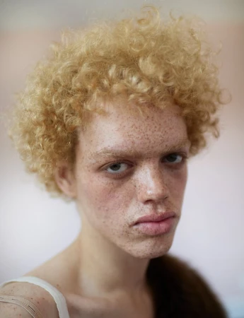 Portrait of a young adult with fair skin and freckles, striking light blonde curly hair, wearing a white tank top and a brown fur stole.