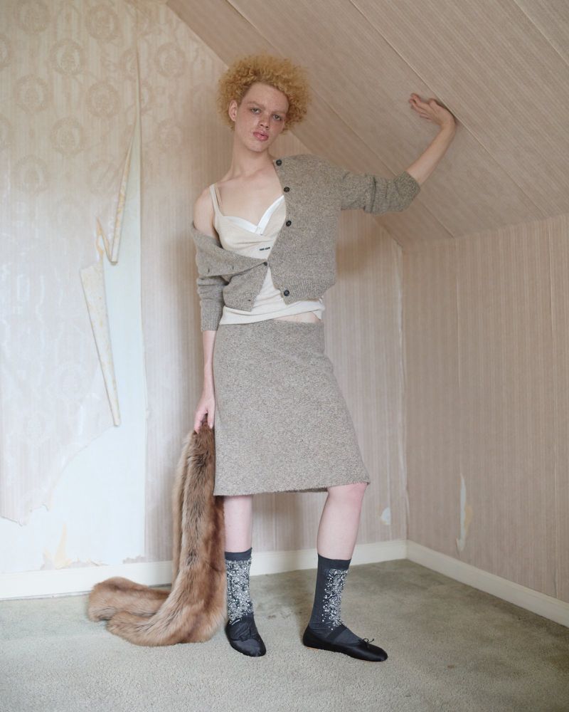 A young adult woman poses in a room wearing a skirt and cardigan, socks, and ballet flats while holding a fox fur stole.