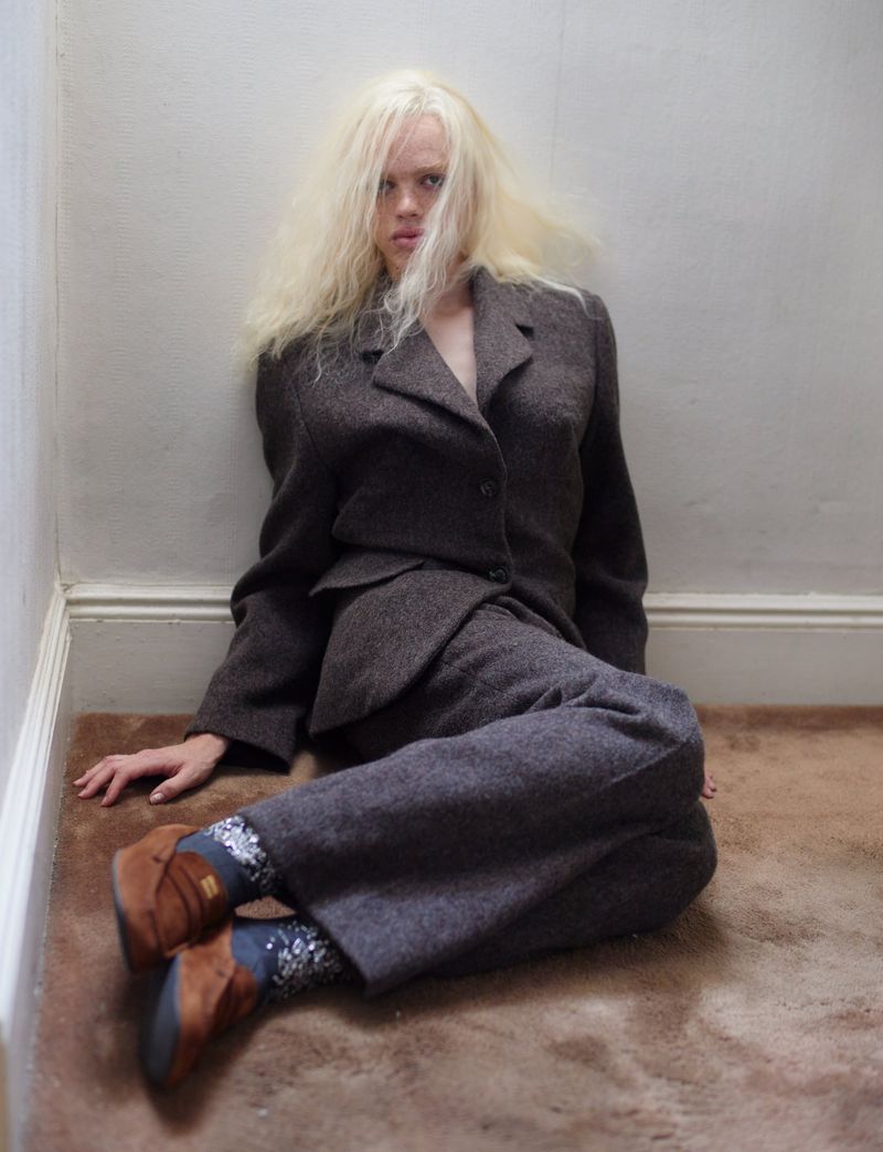 Fashion shot of an adult female with long blonde hair in a brown suit sitting against a wall on a brown carpet, indoors, studio lighting.