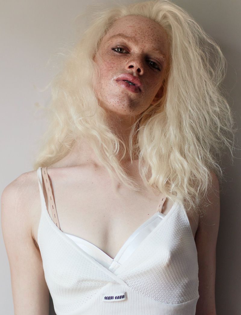 Close-up portrait of a female fashion model with long blonde wavy hair wearing a white spaghetti strap top, posing against a light background.