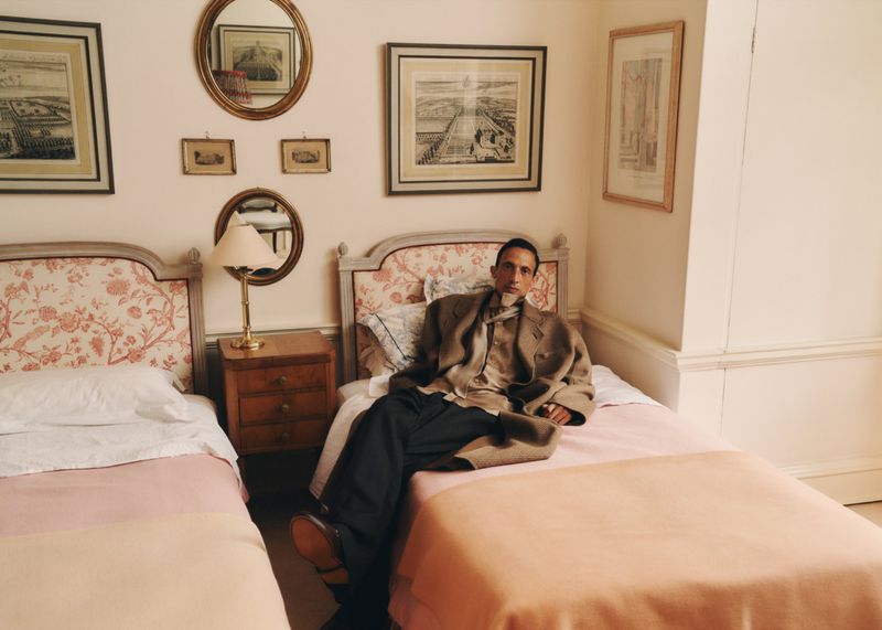 Man in a blazer poses sitting on one of two twin beds in a room with floral headboards, framed art, and mirrors on a light-colored wall.