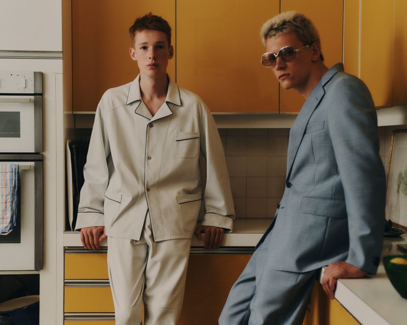 Two fair-skinned young men pose indoors. One wears light blue suit with sunglasses; the other, light pajamas, set against yellow cabinets.