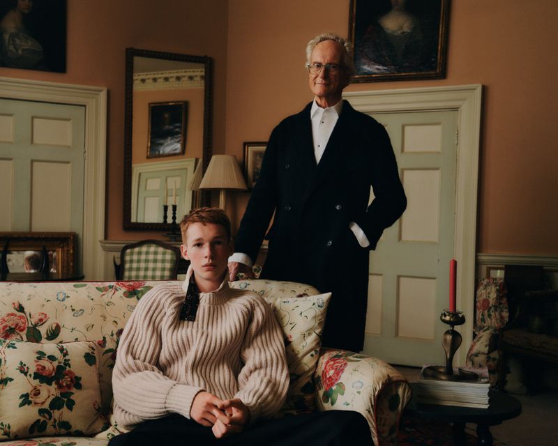 Portrait of a senior man in a black coat with a young, red-haired man on a floral couch in an interior room setting with paintings.