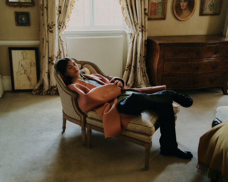 A person lounges in an upholstered armchair in a room with paintings, a wooden dresser, and floral patterned curtains, exuding an editorial fashion vibe.