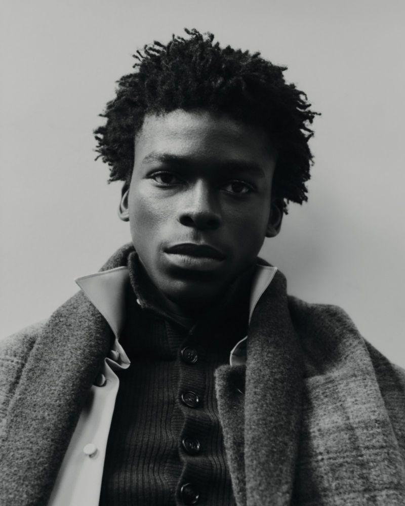 Monochromatic portrait of a Black man wearing a layered plaid coat, knit cardigan, and popped collar shirt against a studio backdrop.
