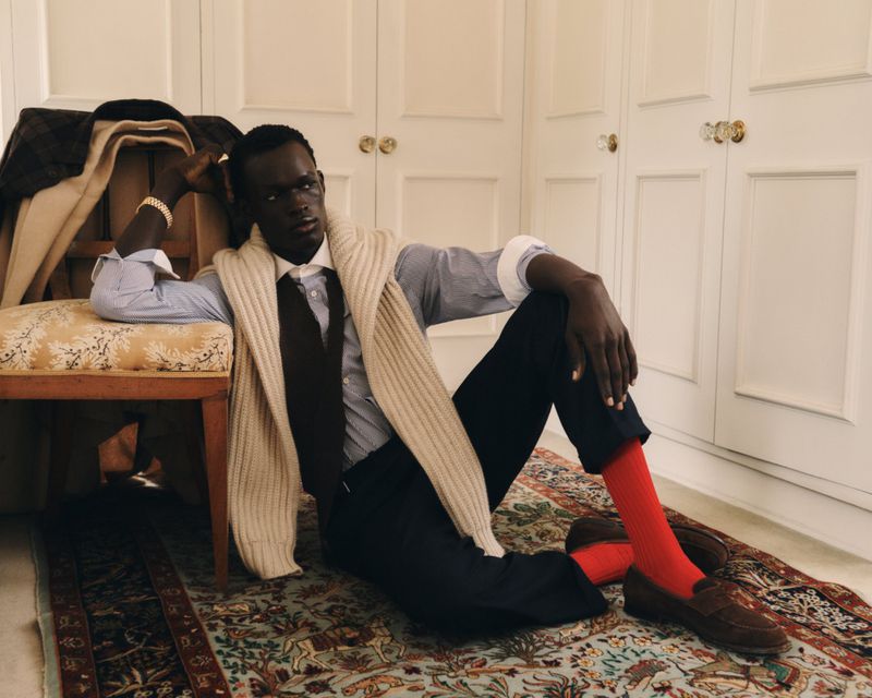 Dark-skinned man lounges indoors in a striped shirt, tie, and red socks, with a beige sweater draped over his shoulders, exuding relaxed style.