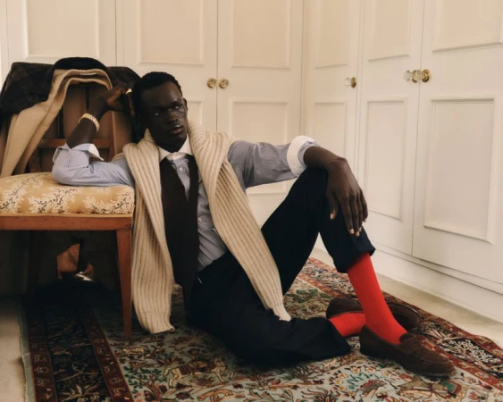 Dark-skinned man lounges indoors in a striped shirt, tie, and red socks, with a beige sweater draped over his shoulders, exuding relaxed style.