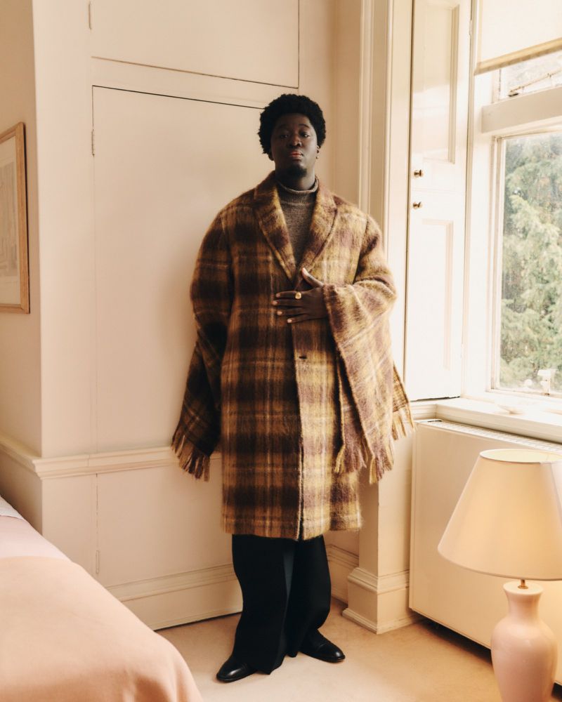 An adult male stands indoors near a window and door. He is wearing a brown and yellow plaid coat with matching scarf and black pants.