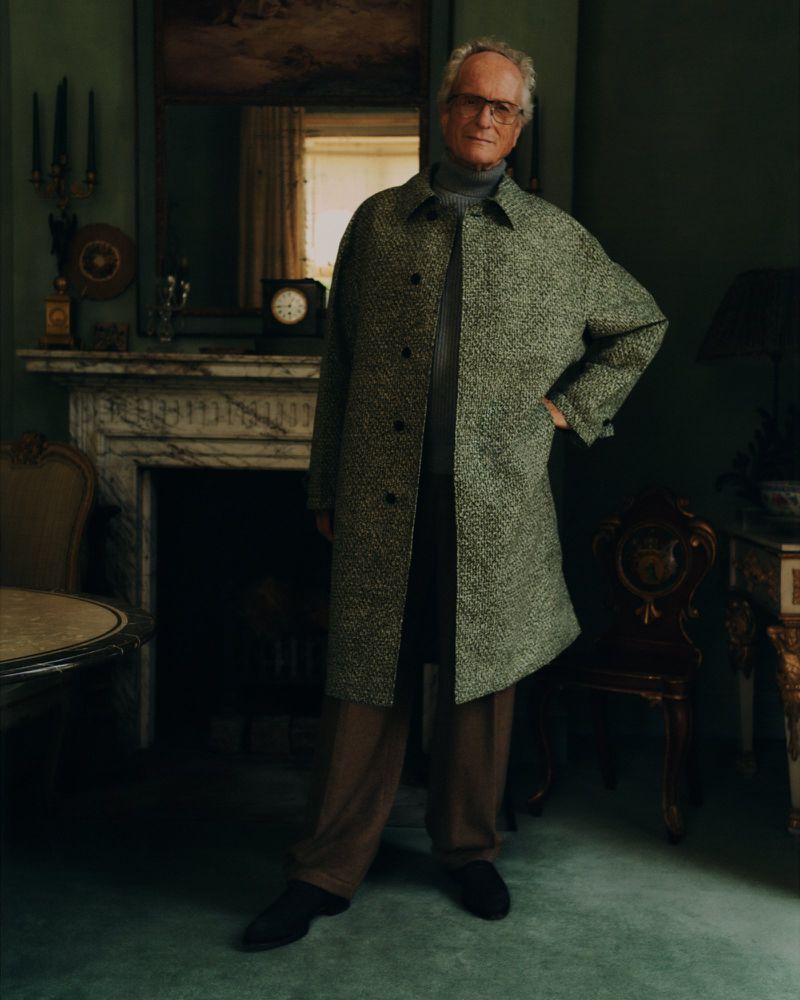 Portrait of an elderly man wearing glasses, a speckled gray coat, and brown pants, standing in a room with a fireplace and ornate decor.