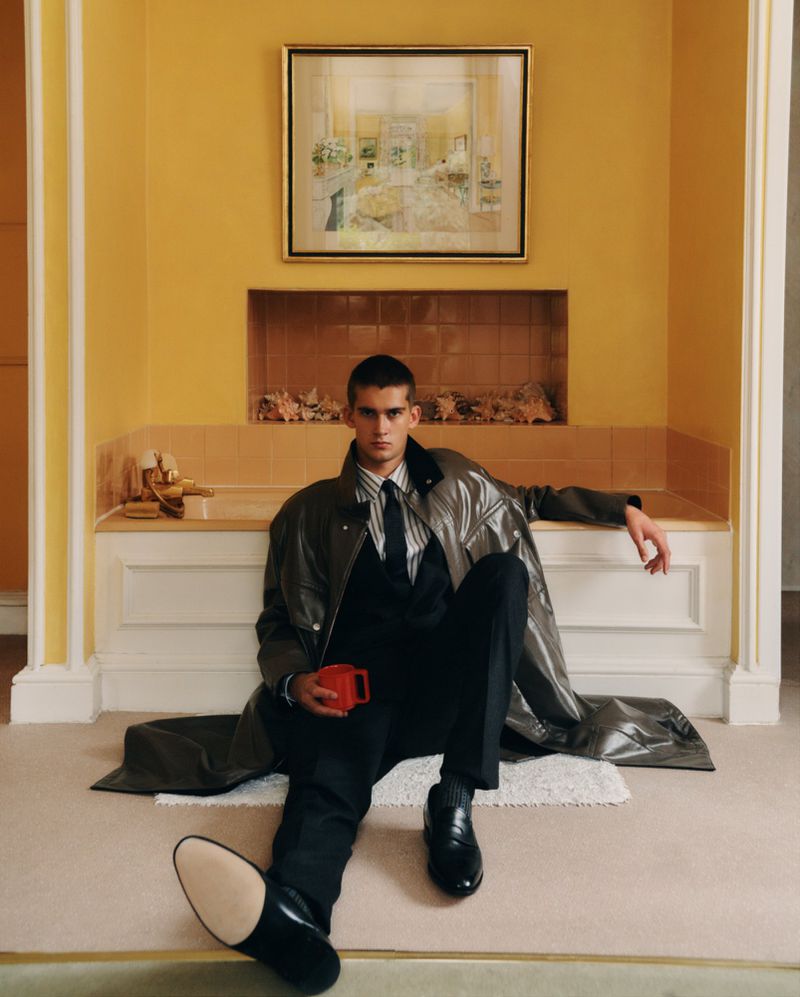 Young man in a dark suit and trench coat sitting on the floor next to a vintage bathtub in front of a yellow tiled wall and painting.