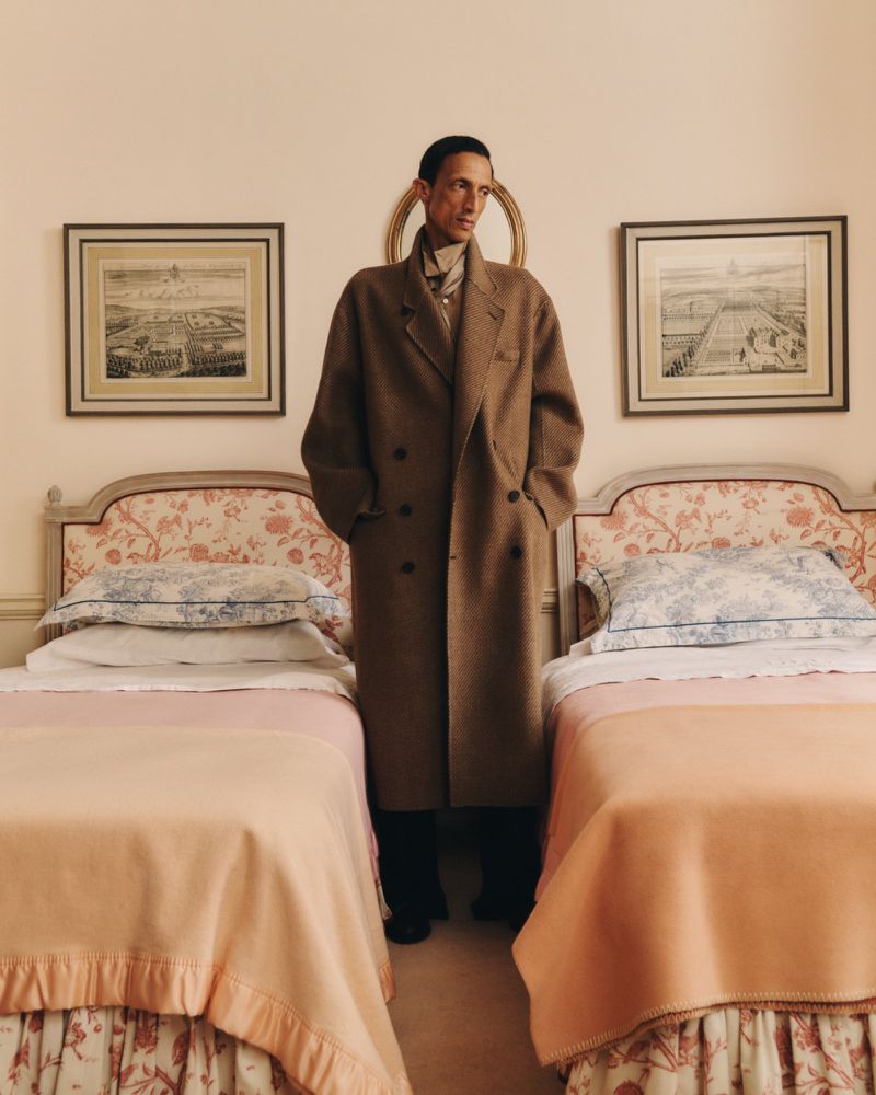 Elegant adult male stands between two twin beds in a light-filled room, wearing a brown wool overcoat, black pants, and a patterned scarf.