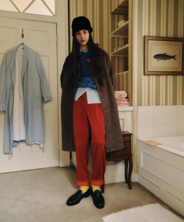 A person in red pants, a brown coat, and a black hat poses in a bathroom with striped wallpaper and an art print of a fish.