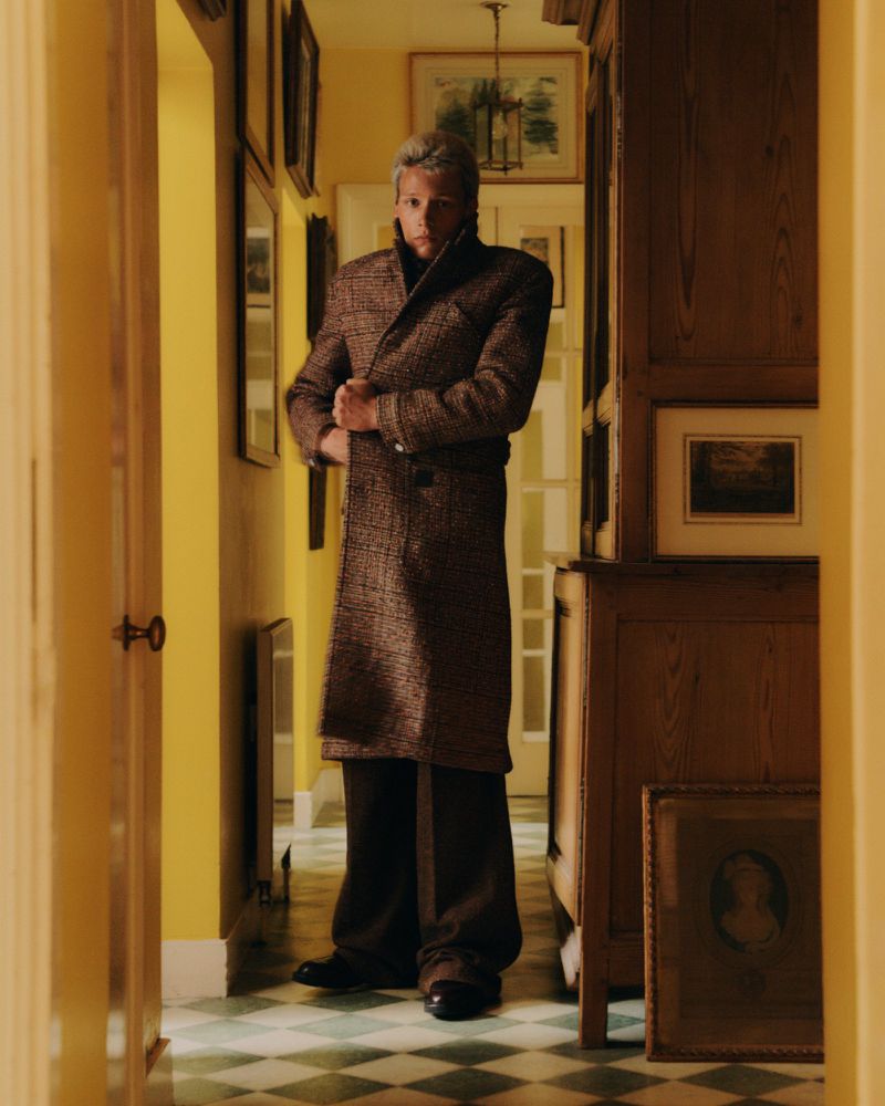 Man stands in hallway wearing a long brown coat, dark pants, and black shoes with a checkered floor, yellow walls, and framed art.