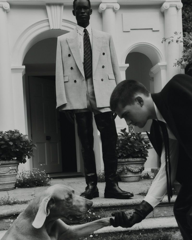 Two men and a dog in front of a building. One man in a suit shakes the dog's paw, another man in a suit stands in the background.