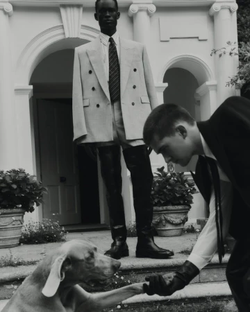 Two men and a dog in front of a building. One man in a suit shakes the dog's paw, another man in a suit stands in the background.