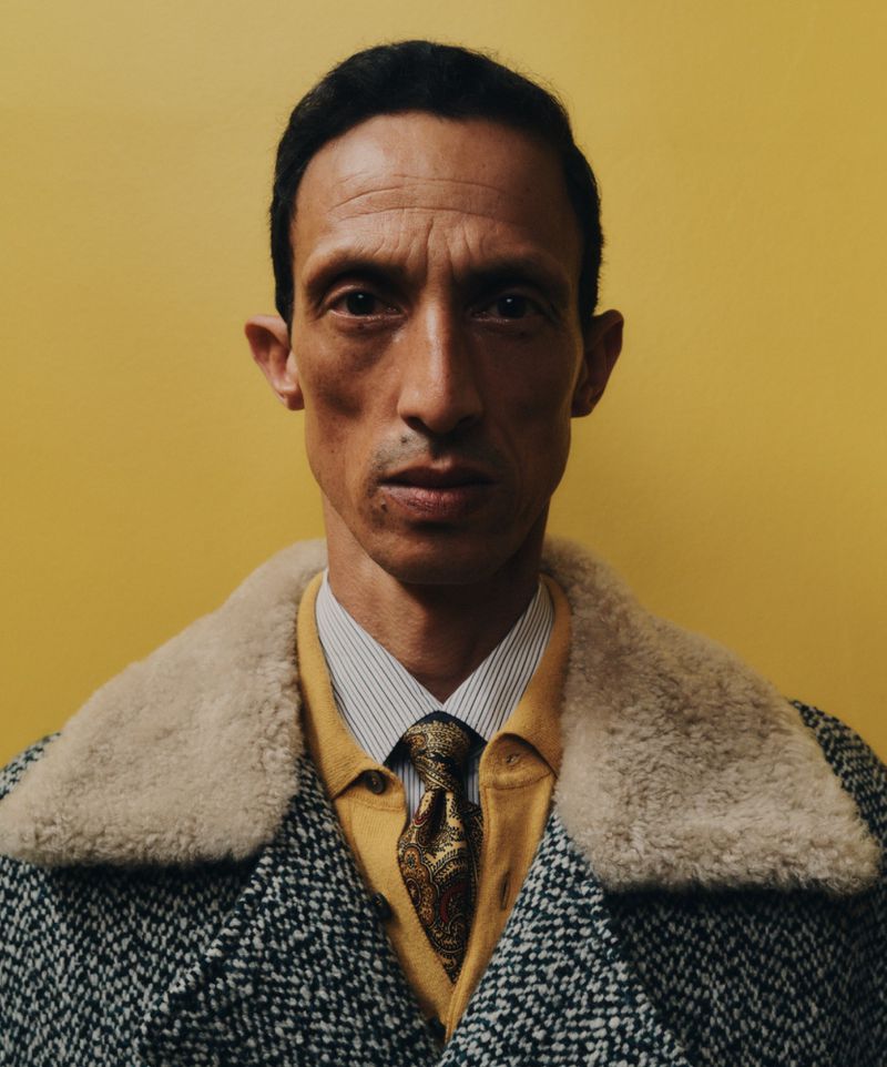 Portrait of a stylish adult male wearing a patterned grey coat with a cream fur collar, yellow sweater, tie, and white striped shirt.