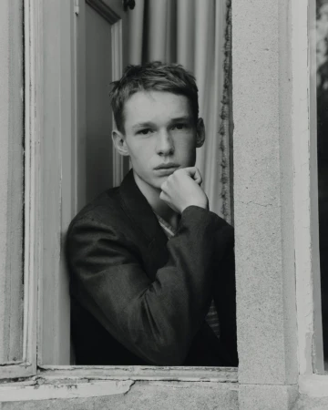 Black and white portrait of a young man resting his chin on his hand while looking out from an open stone window frame.