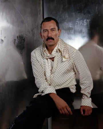 Portrait of an adult male with a mustache wearing a patterned shirt and scarf, sitting against a metallic wall with writing.