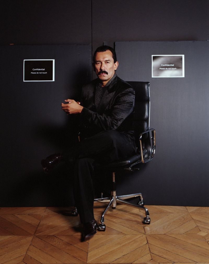 Portrait of an adult male with a mustache in a dark blazer sitting in a black office chair against a dark wall with parquet flooring.