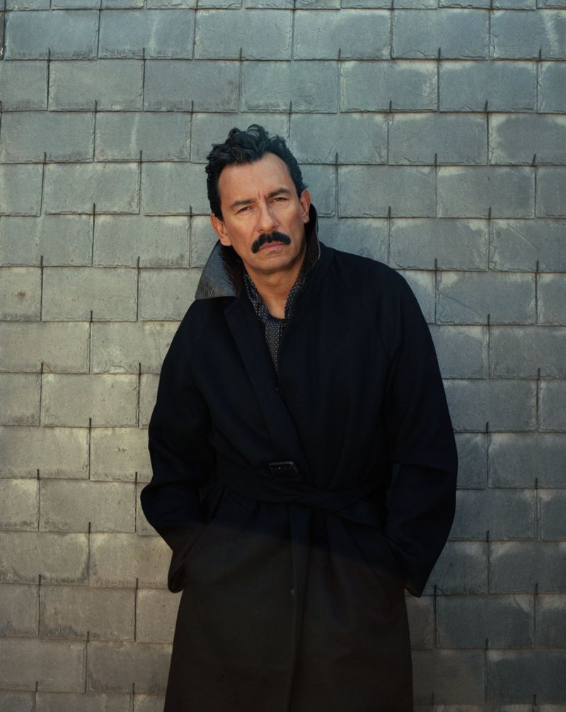 An adult male with a mustache stands in front of a brick wall wearing a dark overcoat and looking off-camera with a serious expression.